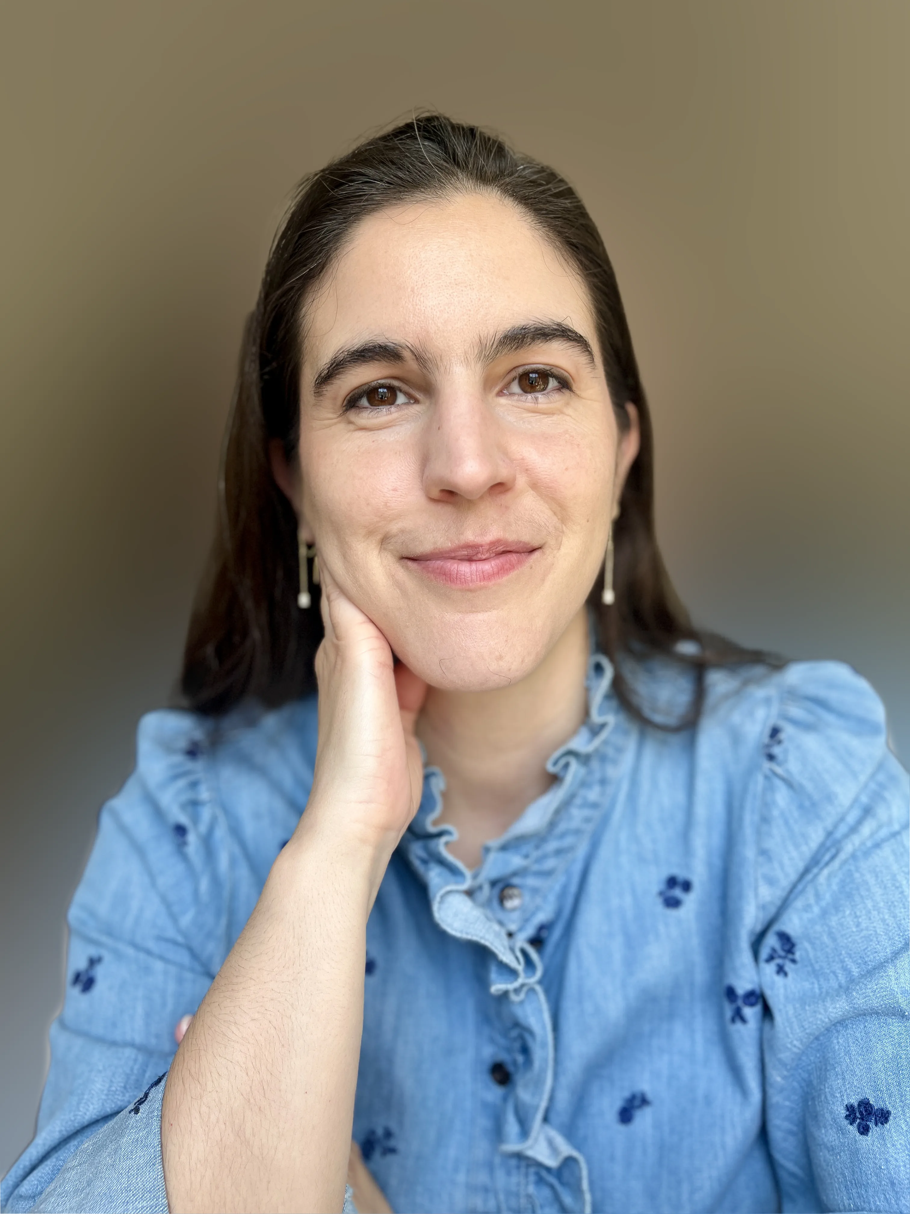 Woman with brown hair smiling while wearing a blue shirt and small white earings